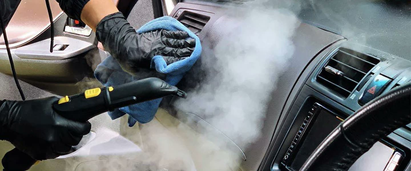 Gloved detailer steam-cleaning a vehicle dashboard with warm mist rising.