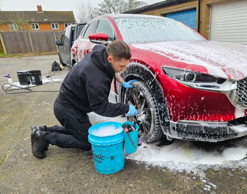 Detailer kneeling beside a foam-covered red Mazda, scrubbing the wheel during a two-bucket wash.