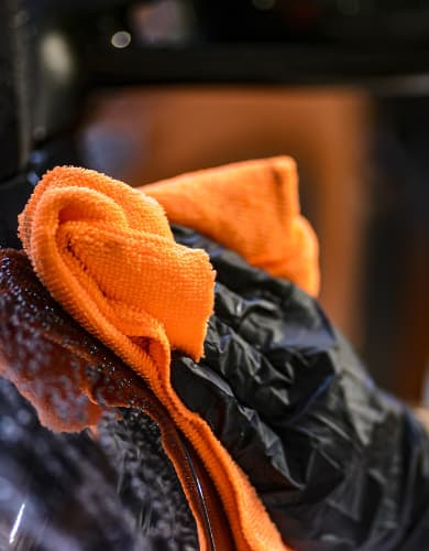 Bright orange microfiber cloth held by a gloved hand, mid-polish on dark paint.