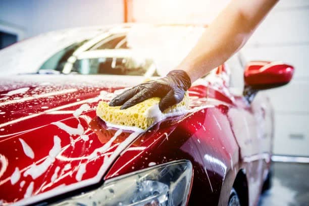 Black-gloved hand with a yellow sponge washing a foamy red car hood, warm sun flare.