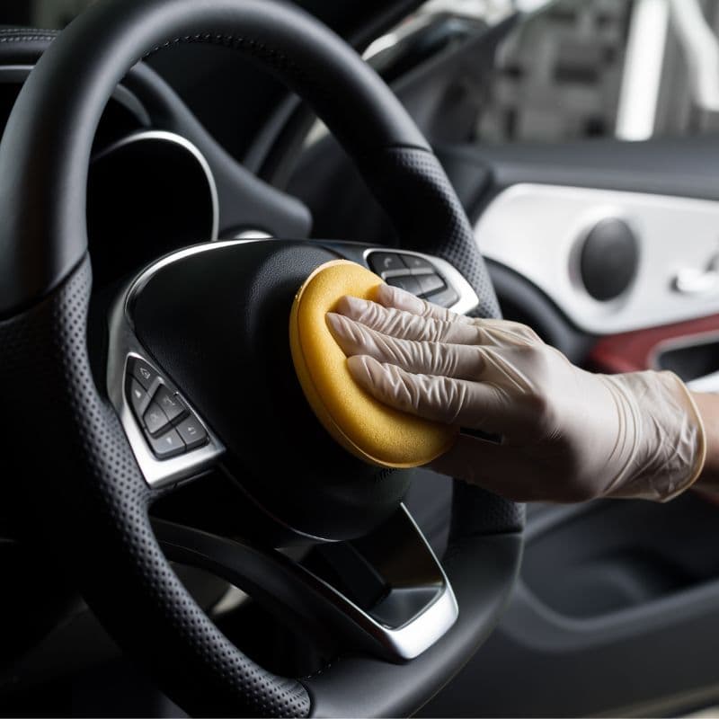 Gloved hand applying conditioner to a leather-wrapped steering wheel.