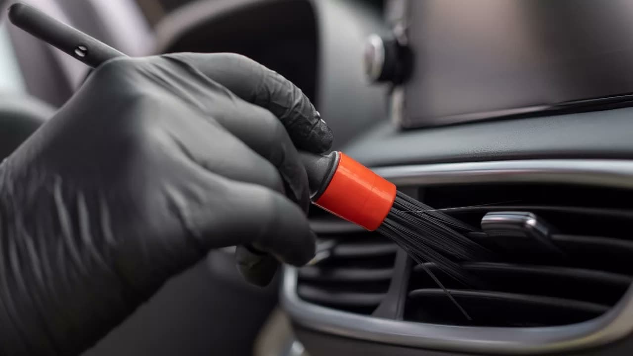 Gloved hand working an orange-handled detail brush into an air vent, dust lifting away.